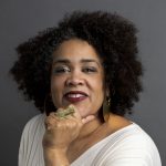 Photo by Margarita Corporan. Professional portrait of Cara, a light-skinned Black woman with an afro, smiling at the camera with a grey backdrop, her chin resting between her closed fist and extended thumb. She is wearing a white sweater, maroon lipstick, and gold earrings, bangles, and a ring.
